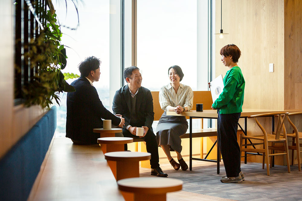 Four people have a casual meeting and conversation in a bright, modern office space with natural light, wooden furniture, and plants visible.