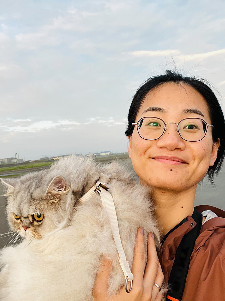 A woman wearing glasses holds a fluffy gray cat outdoors, both facing the camera. The cat appears to be wearing a harness.