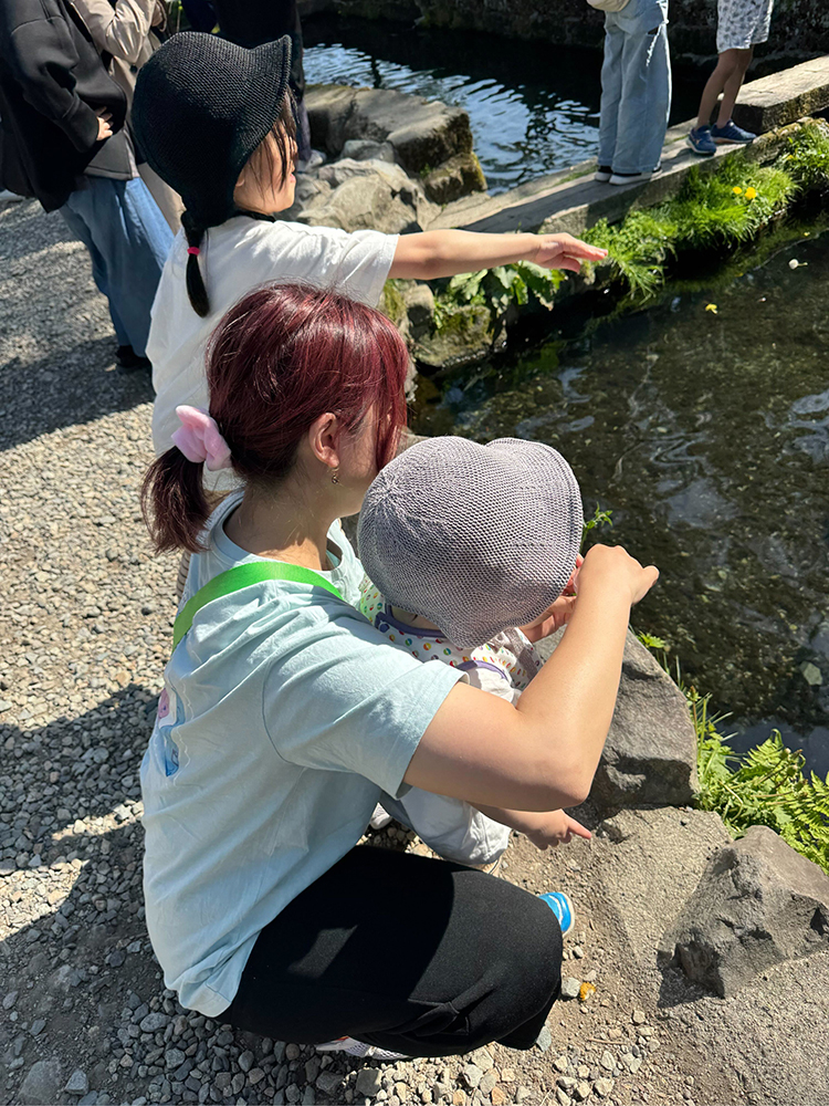 A family enjoying a sunny day by a river, feeding fish together. The scene features adults and a child near the waterside, surrounded by rocks and greenery.