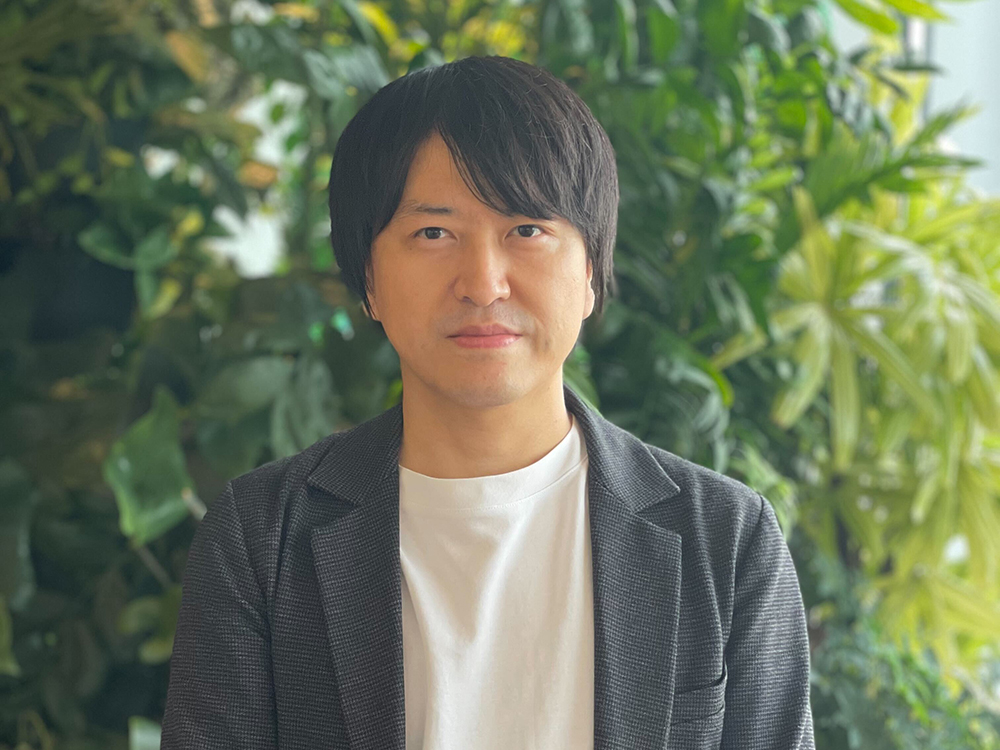 A man wearing a dark blazer and white shirt standing in front of a lush green background with various plants.