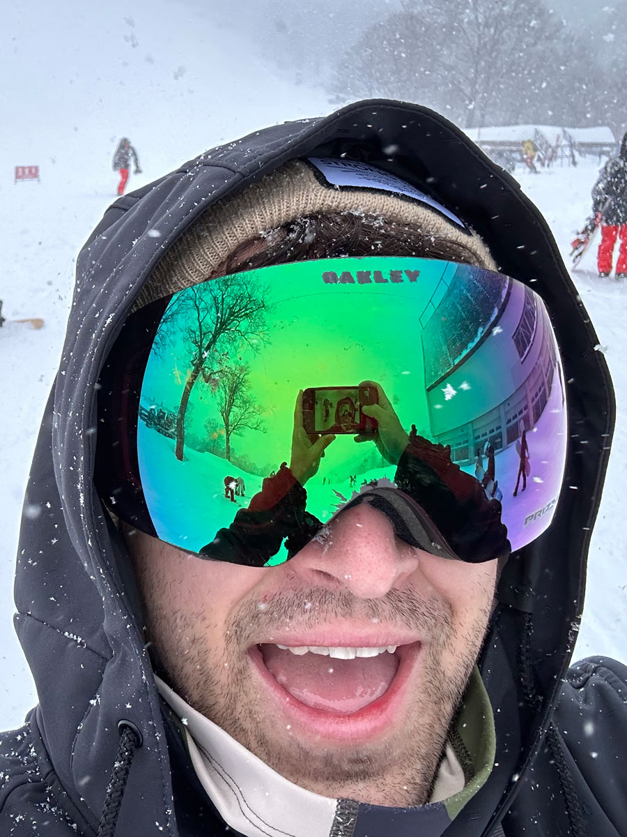 A close-up selfie of a person wearing reflective Oakley goggles on a snowy mountain. The reflection in the goggles shows the snowy landscape and the person taking the photo.