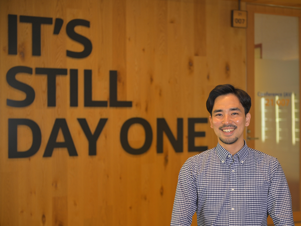 A man smiles while standing in front of a wooden wall with a large sign reading 'IT'S STILL DAY ONE.'