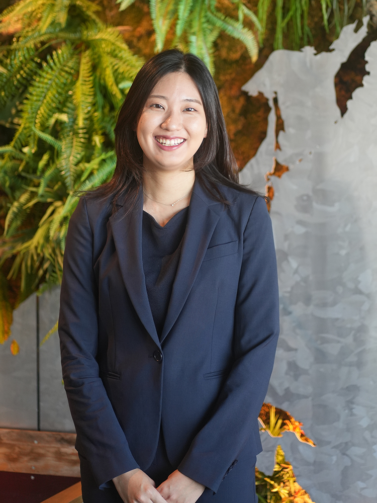 A businesswoman in a navy suit smiling while standing indoors, with lush green plants and modern decor in the background.