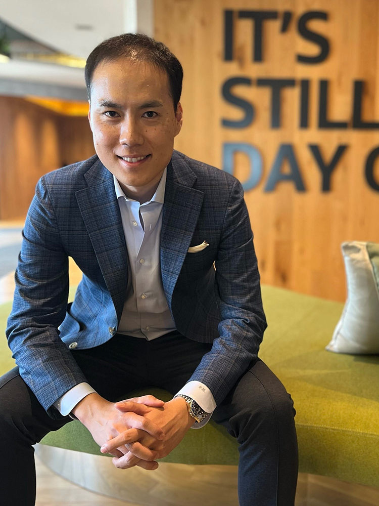 A businessman in a blue suit sits on a green couch in a modern office setting, with a wall sign in the background reading 'IT'S STILL DAY ONE.'