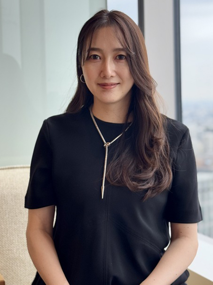 A woman with long brown hair wearing a black dress, sitting and smiling in a modern office setting with large windows in the background.