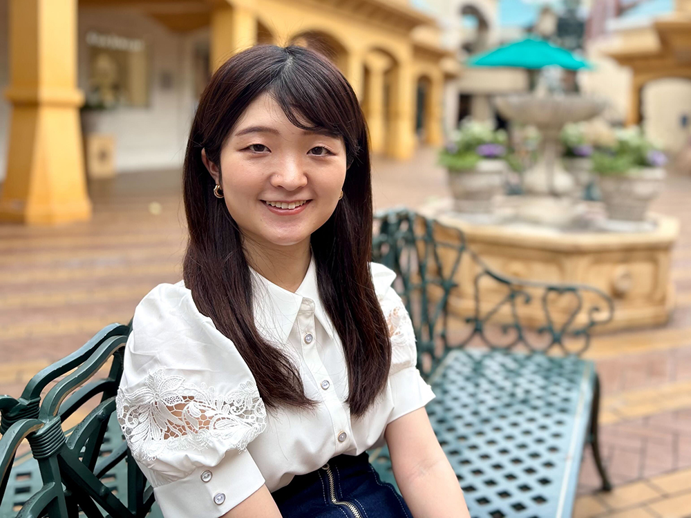 A woman smiling while sitting on a decorative metal bench in an outdoor plaza, with a fountain and plants in the background.