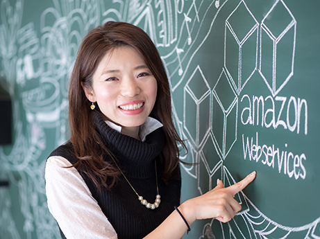 A woman smiling and pointing at a chalkboard drawing featuring the Amazon Web Services (AWS) logo and text.