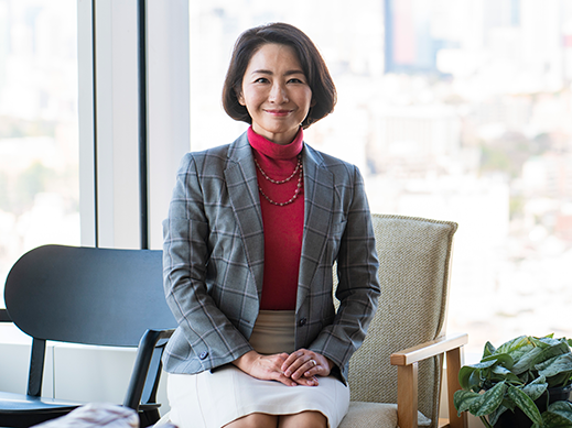 A businesswoman sits in an office chair in front of a large window with natural light, dressed in a plaid blazer, red turtleneck, and white skirt, smiling at the camera. The modern office features cityscape views and indoor plants.