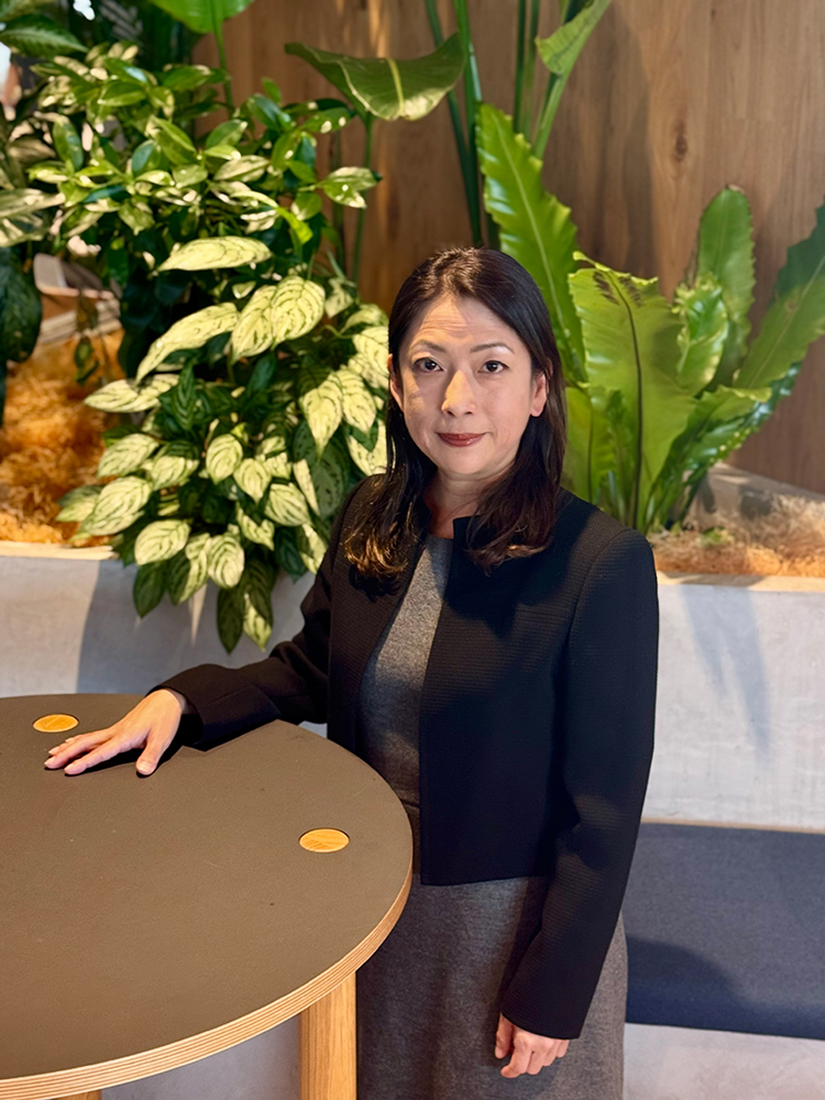 A woman stands next to a round table surrounded by lush indoor plants in a modern office setting.