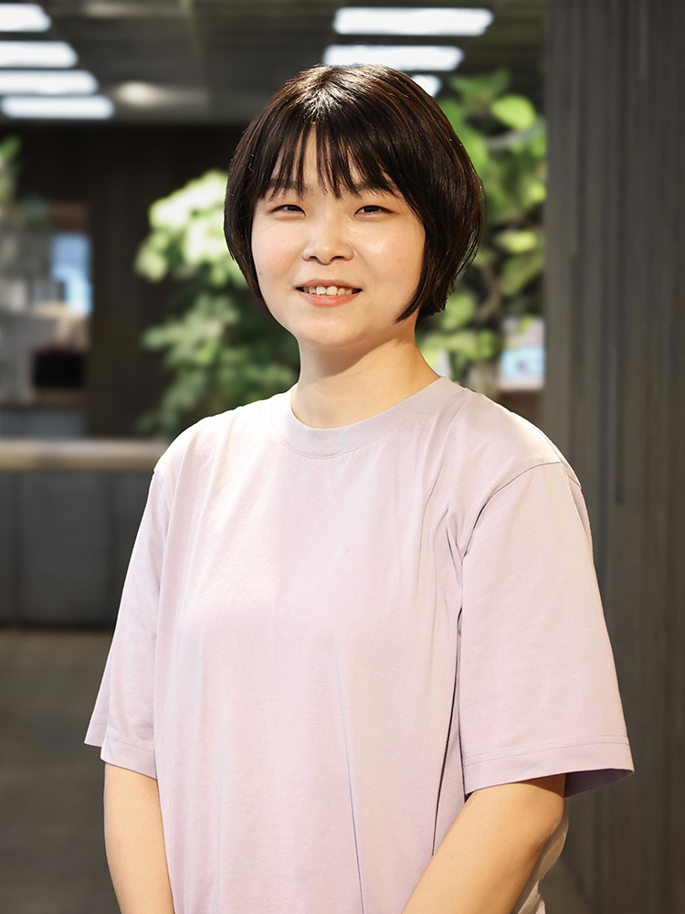 A woman wearing a light purple shirt, standing indoors with a softly lit background and greenery visible. Portrait orientation.