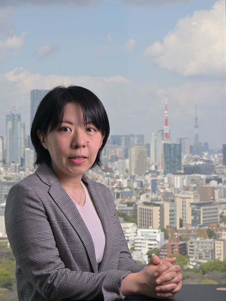A businesswoman in a plaid blazer sits in front of a large window with the Tokyo city skyline, including Tokyo Tower and Skytree, visible in the background.