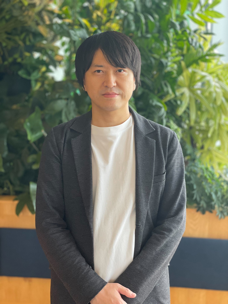 A man wearing a dark blazer and white shirt stands indoors in front of a background of lush green plants.