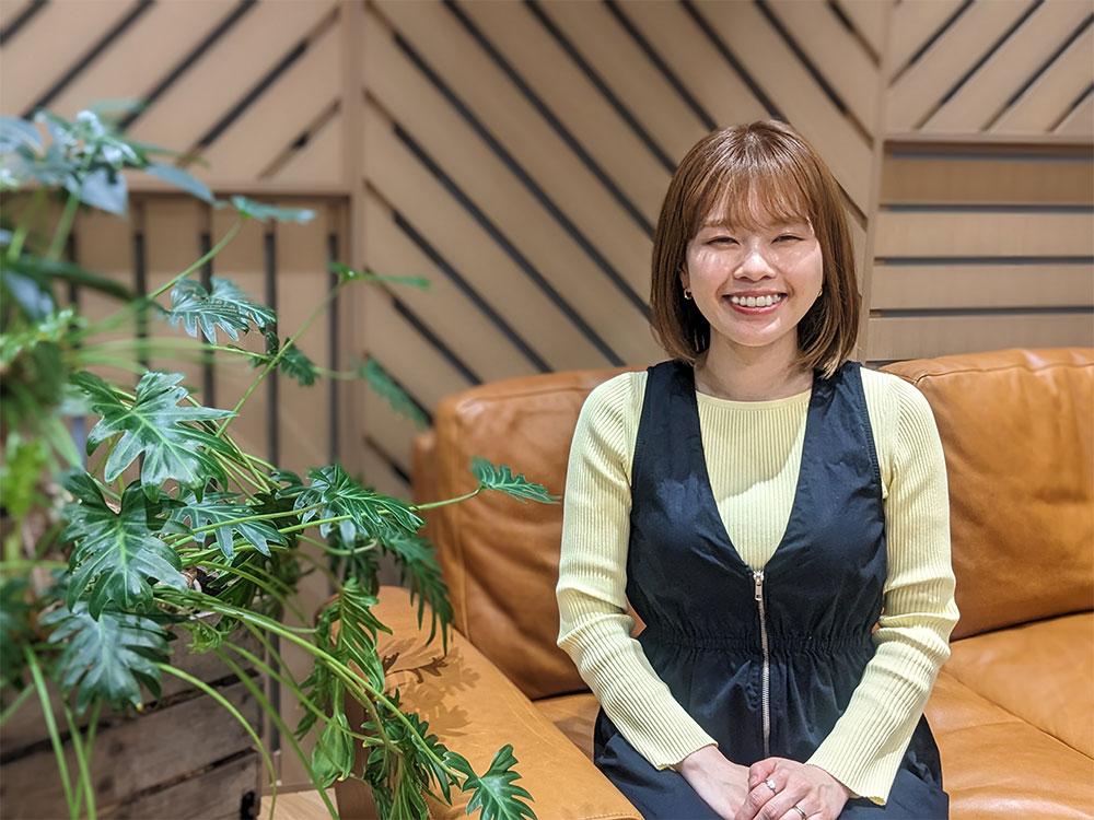 A woman smiling while sitting on a brown couch next to a green plant. The background features a modern wooden wall design.