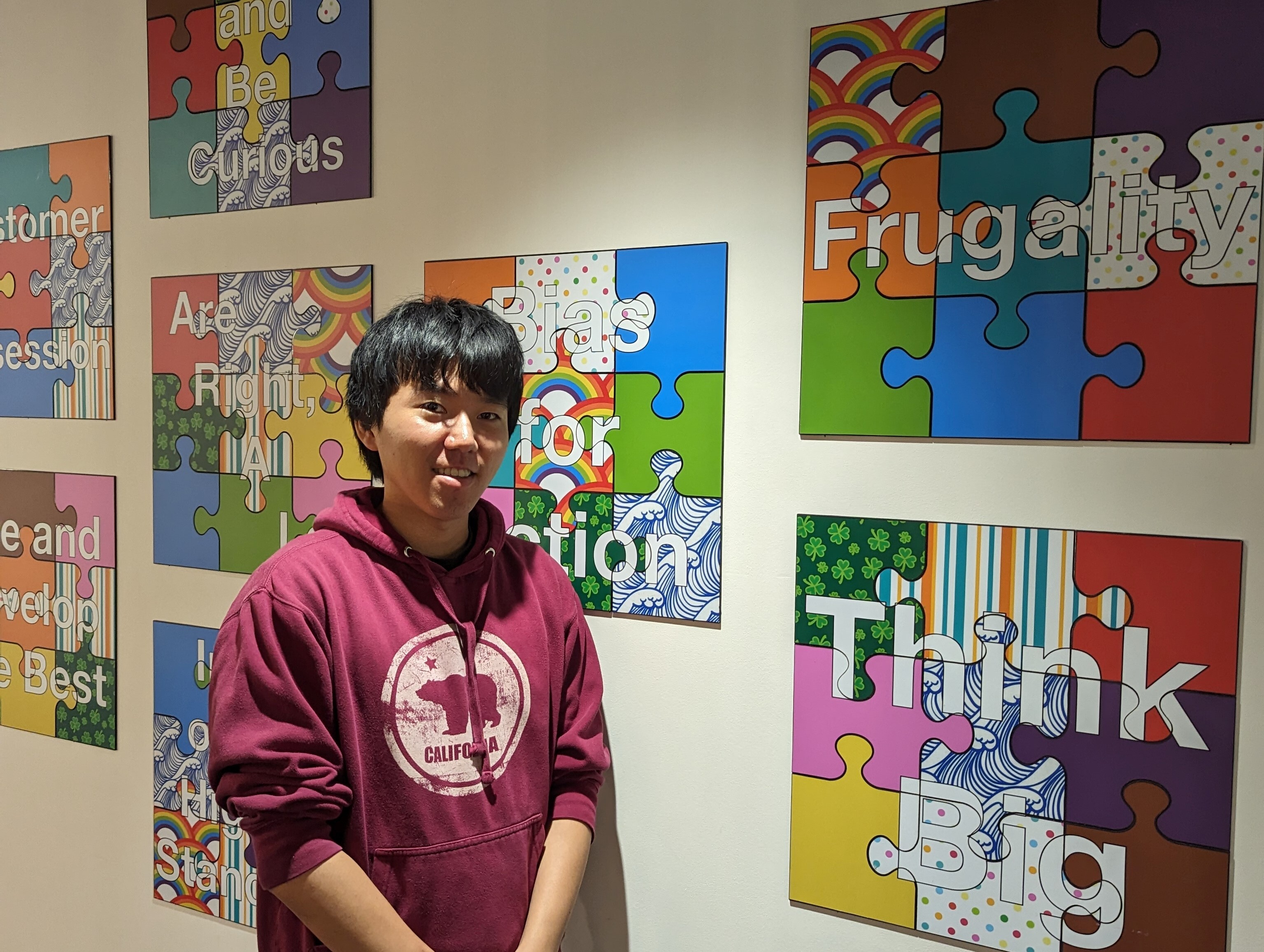A person wearing a maroon hoodie stands in front of colorful wall art made of puzzle pieces with motivational words and phrases such as 'Frugality' and 'Think Big'.