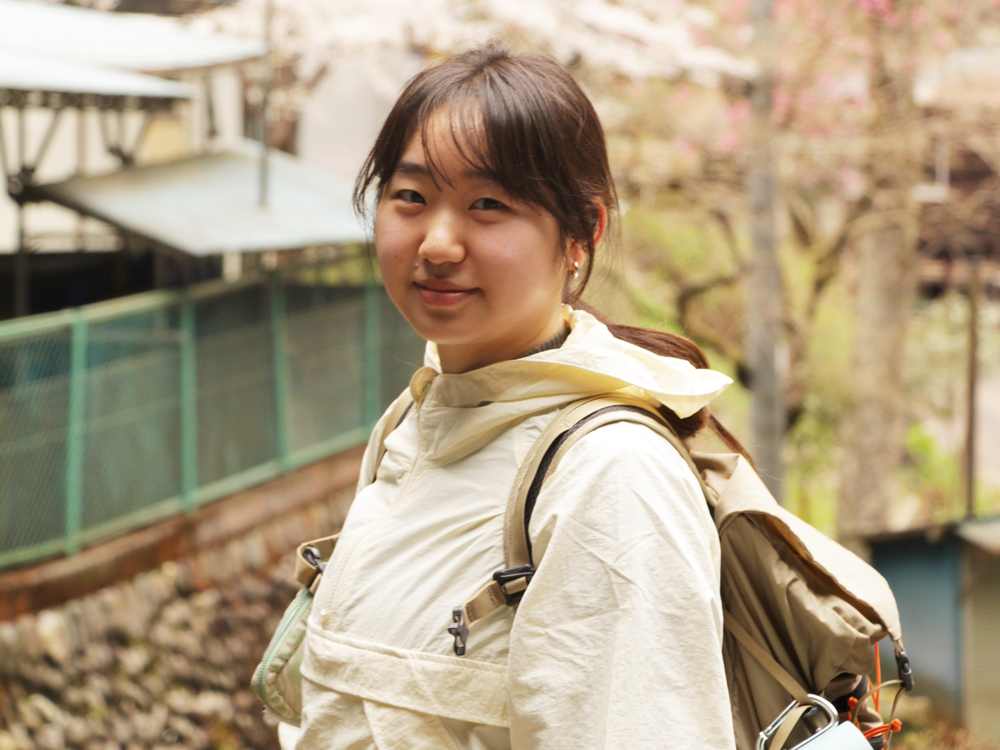 A young woman wearing a light jacket and a backpack, outdoors in a natural setting, possibly on a hike.