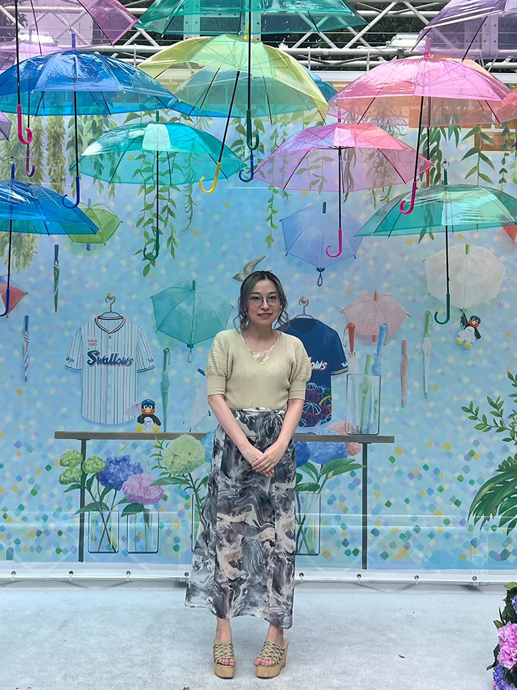 A woman stands beneath a display of colorful hanging umbrellas with a decorated background featuring flowers, sports jerseys, and plants.