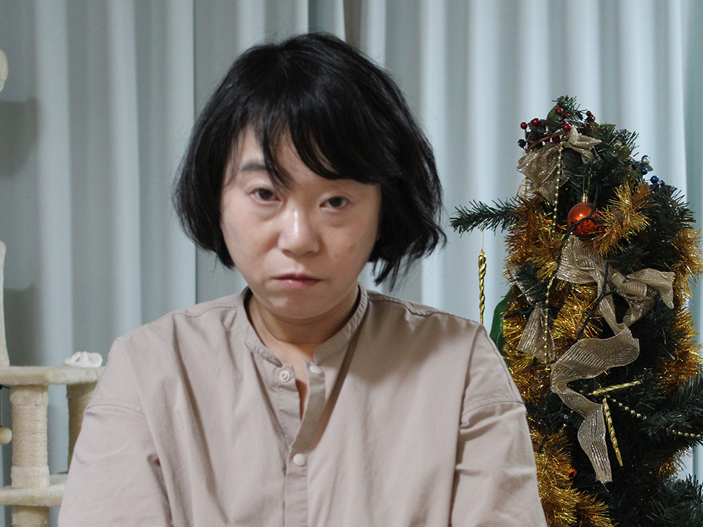 A woman with a neutral expression is sitting indoors with a decorated Christmas tree in the background.