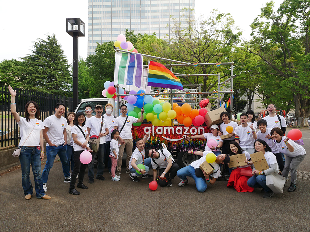 A group of Amazon employees participating in a pride parade, gathered in front of a decorated float with rainbow flags and colorful balloons. The team is wearing white shirts with rainbow heart logos and holding balloons, celebrating diversity and inclusion.
