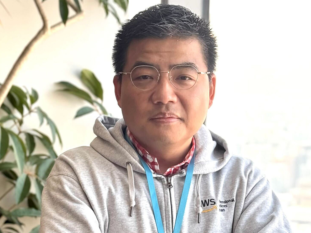 Man wearing AWS hoodie and glasses in an indoor portrait setting, with plants and a window in the background.