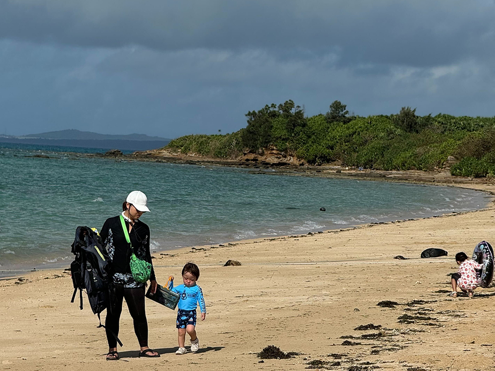 A family spending time together on a secluded sandy beach with blue water, greenery in the background, and children playing near the shore.