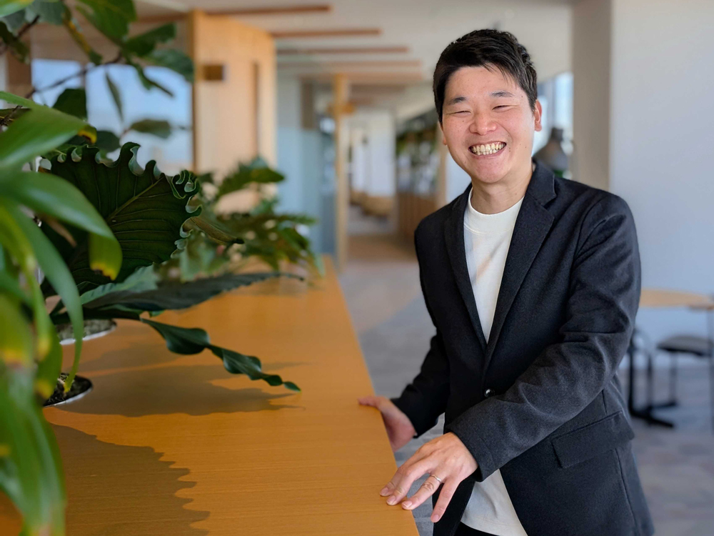 A smiling man wearing a dark blazer is standing at a wooden counter in a modern office space decorated with green plants.