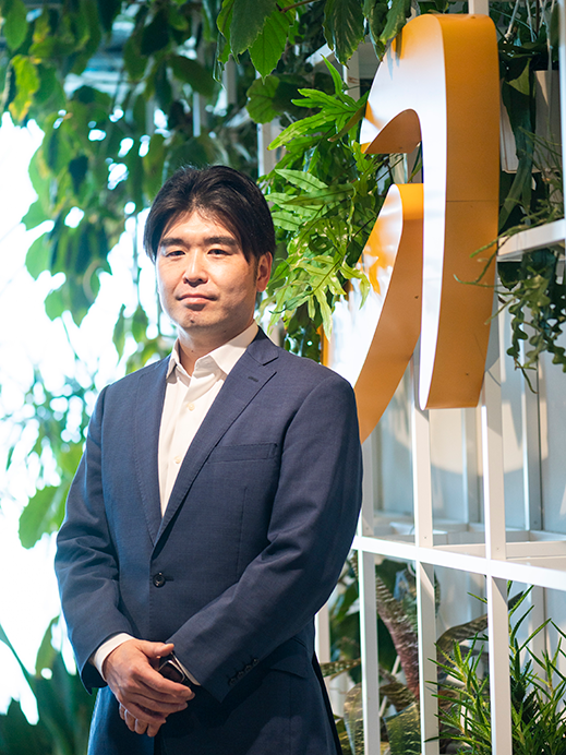 A businessman in a suit stands in front of a large Amazon logo surrounded by indoor plants.