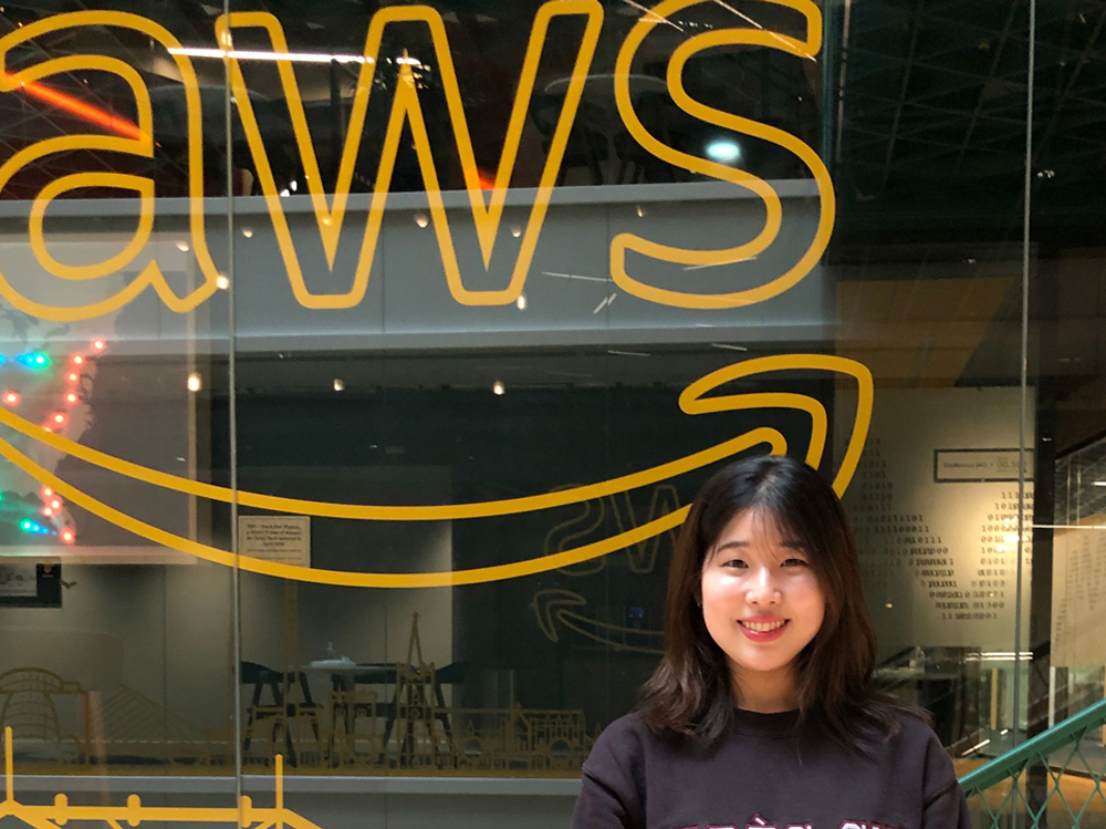 A woman smiling while standing in front of a large glass wall with the yellow AWS logo. The setting appears to be an AWS office or building.