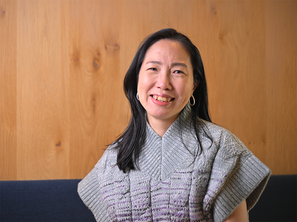 A woman smiling while wearing a gray sweater, seated in front of a wooden background.