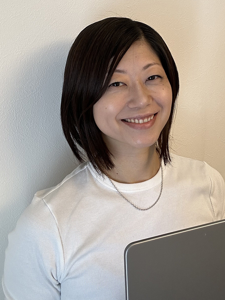 A woman smiling while holding a laptop and wearing a white shirt, standing against a light-colored wall.