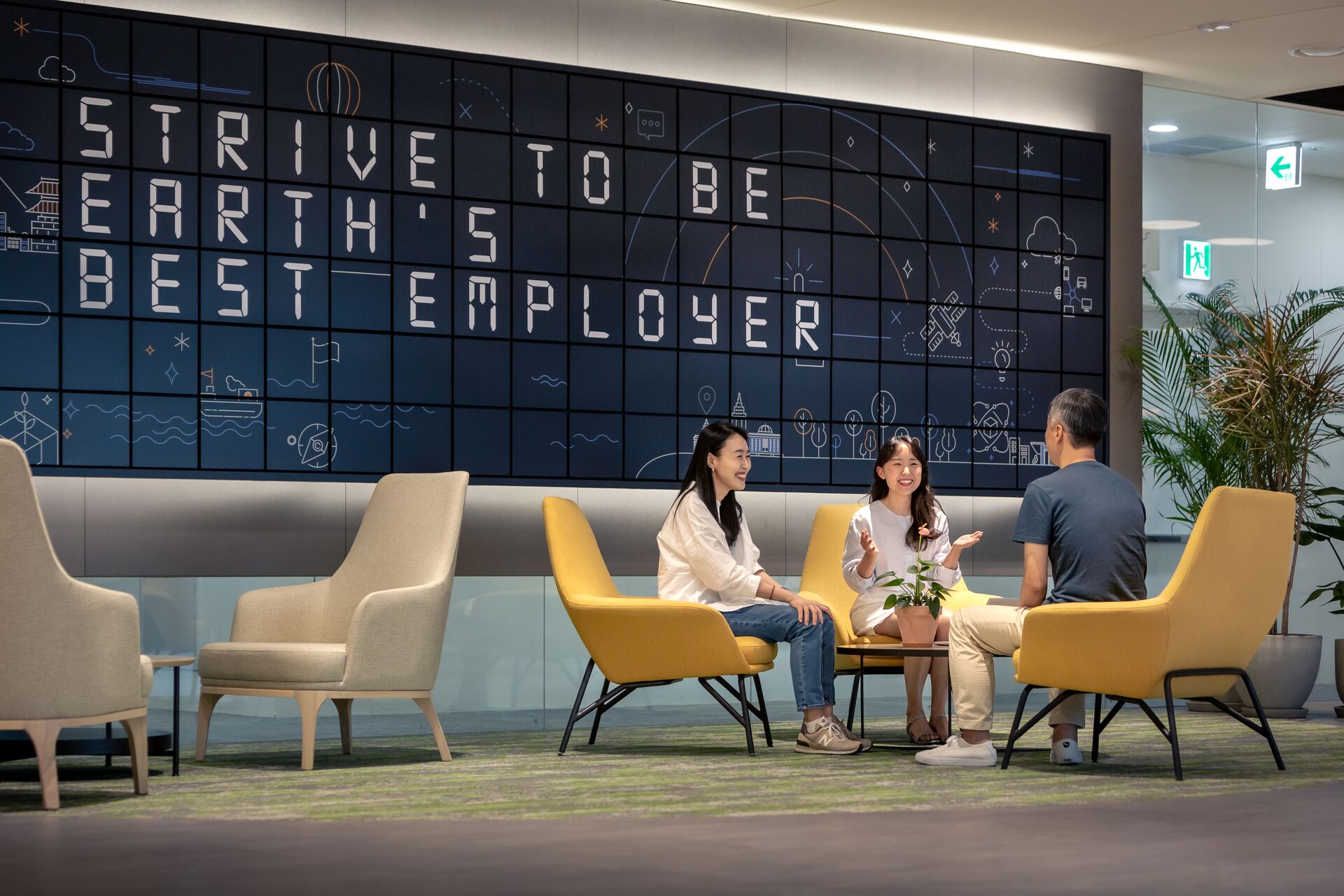 Three people sitting in a modern office lounge area with yellow chairs, a small plant on the table, and a wall display reading "Strive to be Earth's Best Employer."