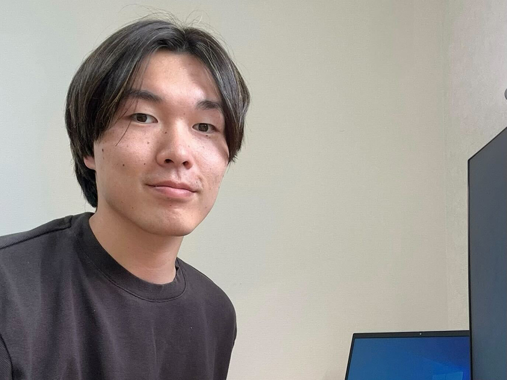 A young man sitting at a desk with a computer monitor in an office or home workspace.