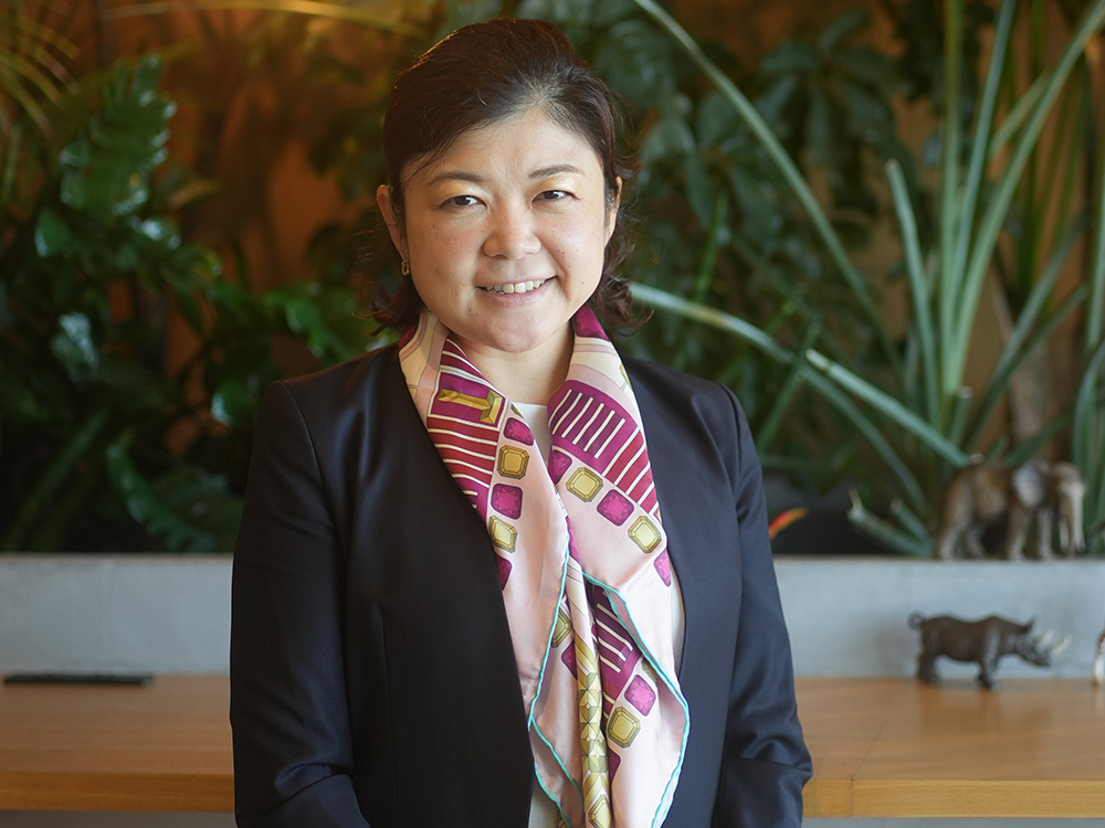 A woman in business attire with a patterned scarf stands in front of a table, with various indoor plants and plant decorations in the background.