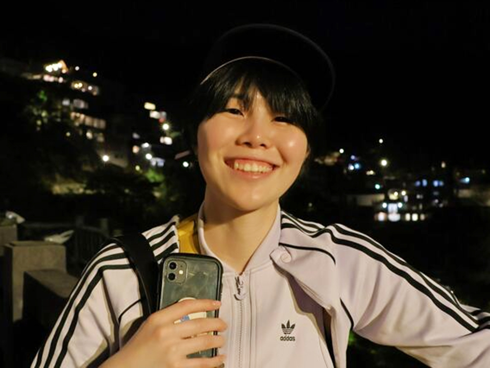 A person smiling and wearing an Adidas jacket and cap, holding a smartphone, with city lights in the background at night.