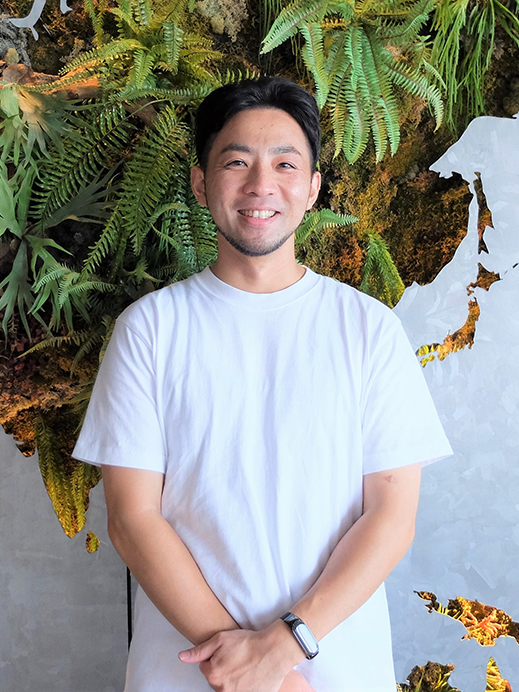 A man wearing a white shirt stands smiling in front of a green plant wall background.