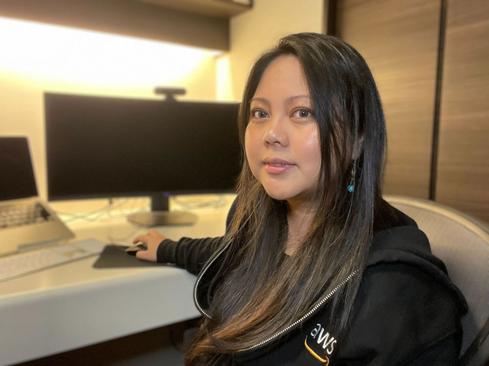 A woman sits at a desk with a computer, wearing an AWS-branded hoodie.
