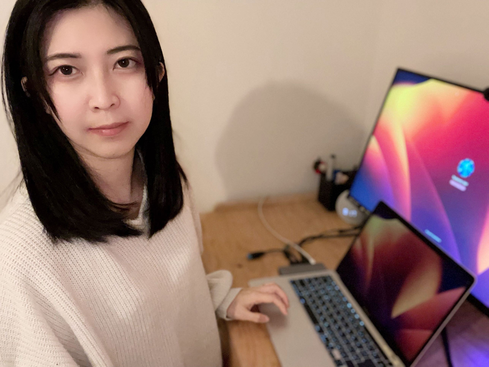 A person working at a desk with a laptop and an external monitor, in a home or office setting.