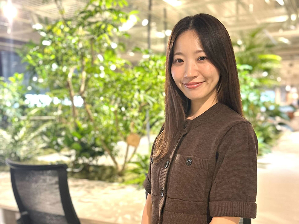 A woman smiling in a modern office setting with lush indoor plants in the background.