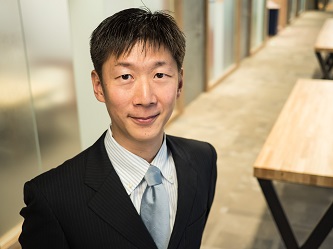 A professional business portrait of a man in a suit standing in a modern office hallway with wooden tables.
