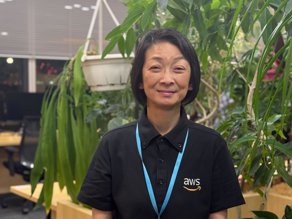An AWS employee stands in an office surrounded by green plants, wearing a black AWS polo shirt and a blue lanyard. The background features office desks, chairs, and large leafy plants, creating a welcoming and natural work environment.