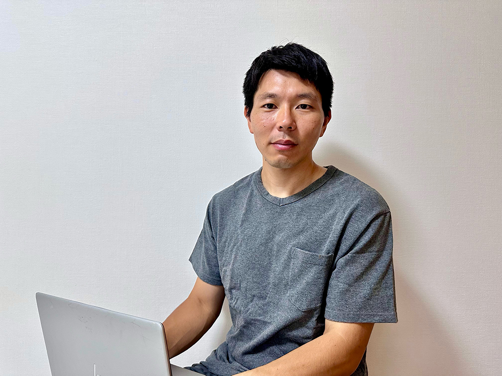 A man in a grey t-shirt sitting and using a laptop against a plain light-colored background.