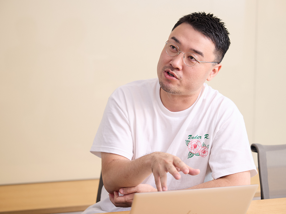A man wearing a white t-shirt and glasses is talking during a meeting, seated at a table with a laptop in front of him.