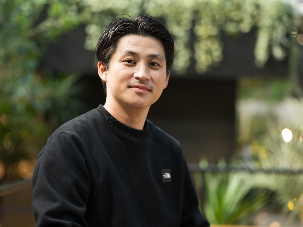 A person wearing a black sweatshirt with a blurred background of greenery and soft lighting.