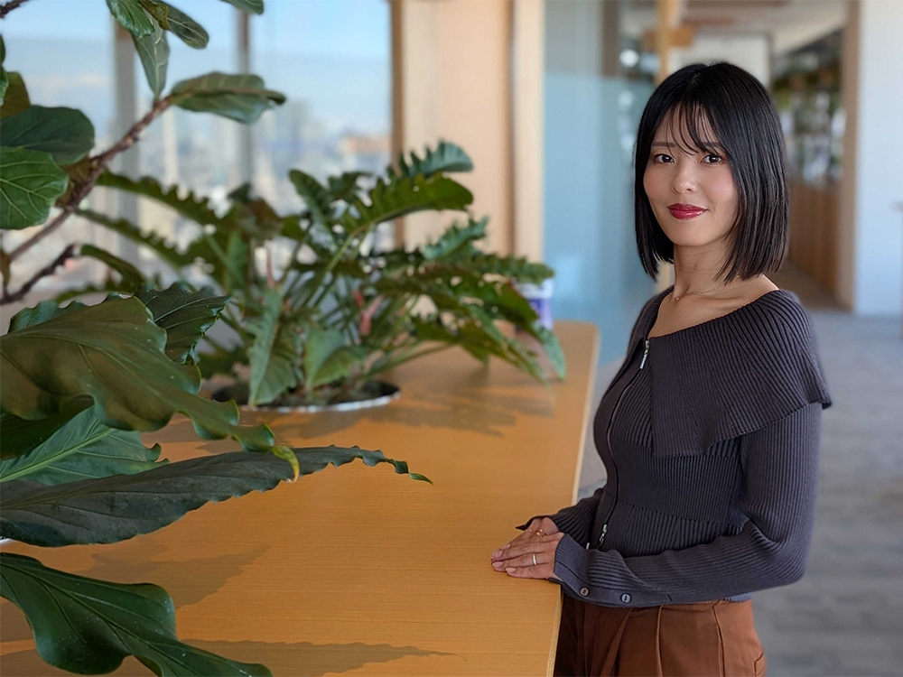 A woman standing in a modern office setting, next to a counter with large green plants in the background.