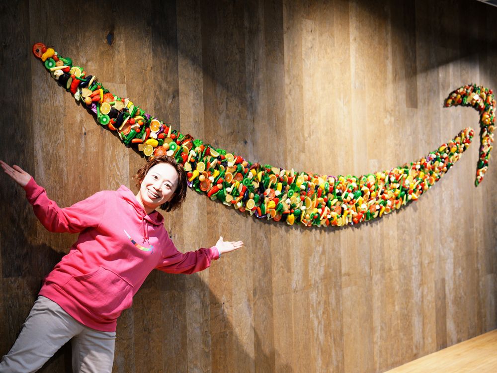 A person wearing a pink hoodie stands in front of a wall decorated with art made from colorful vegetables arranged in the shape of the Amazon logo.