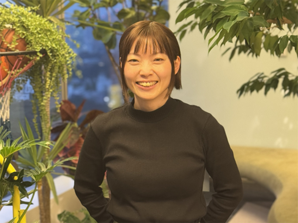 A woman smiling while standing indoors, surrounded by various green plants and foliage in the background.