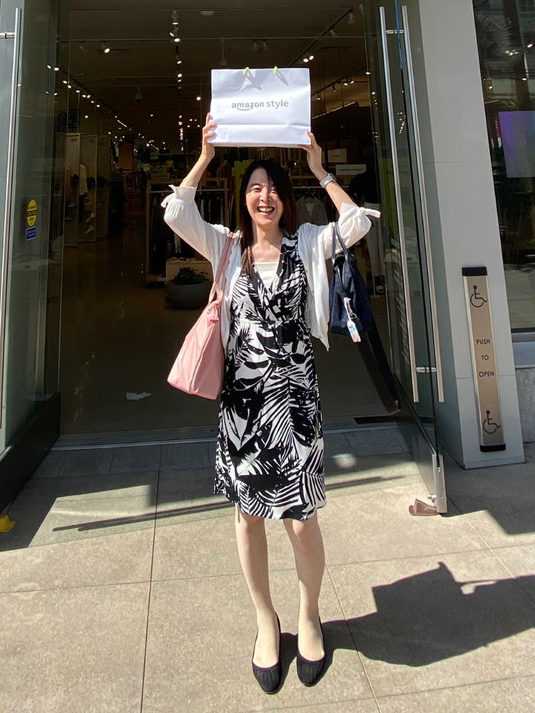 A woman stands outside a store entrance holding a shopping bag labeled 'amazon style' above her head and smiling in the sunlight.