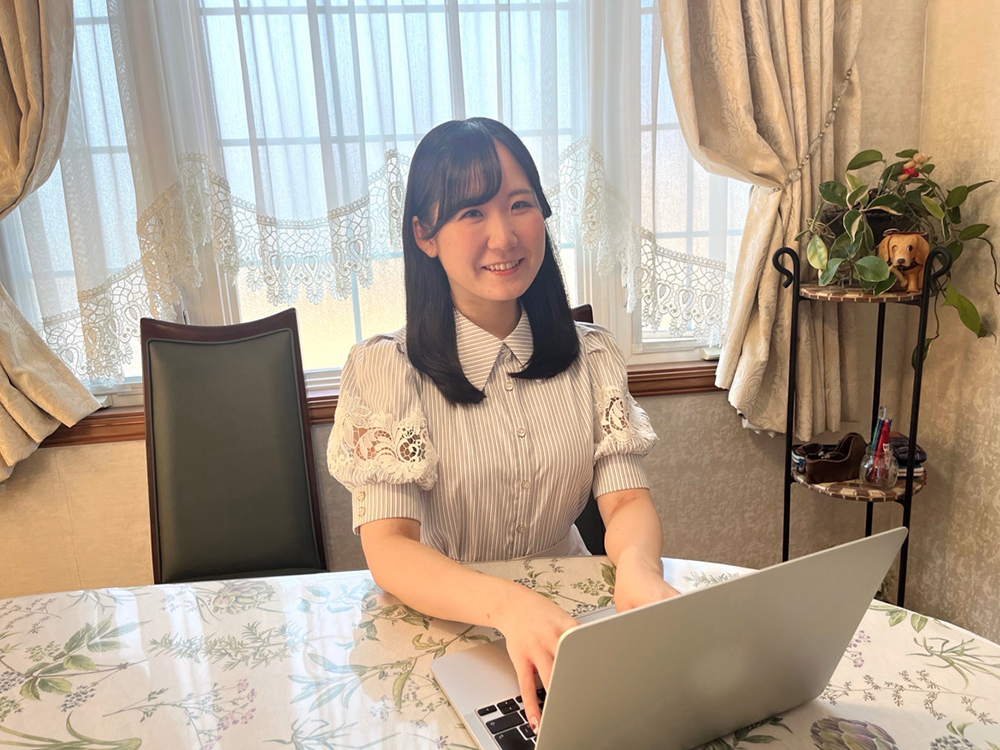 A woman is smiling while using a laptop at home, sitting at a table with a floral tablecloth in a bright, cozy room with lace curtains and indoor plants.