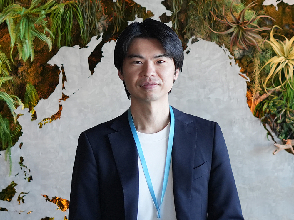 A man wearing a suit and a blue lanyard stands in front of a green wall background with plants and foliage.