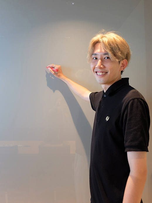 A smiling person with blonde hair is writing on a whiteboard with a marker while looking at the camera.