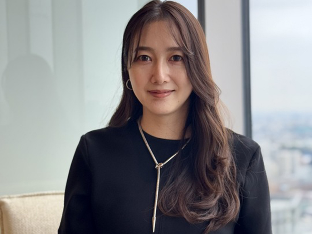 A woman with long hair wearing a black top is seated in front of a window with a city view in the background.